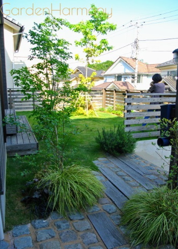 庭の眺め（玄関アプローチから）：天然木のウッドフェンスに囲まれた庭は、過不足ないシンプルな印象ながら、植物の緑がみずみずしいナチュラルな庭です。
